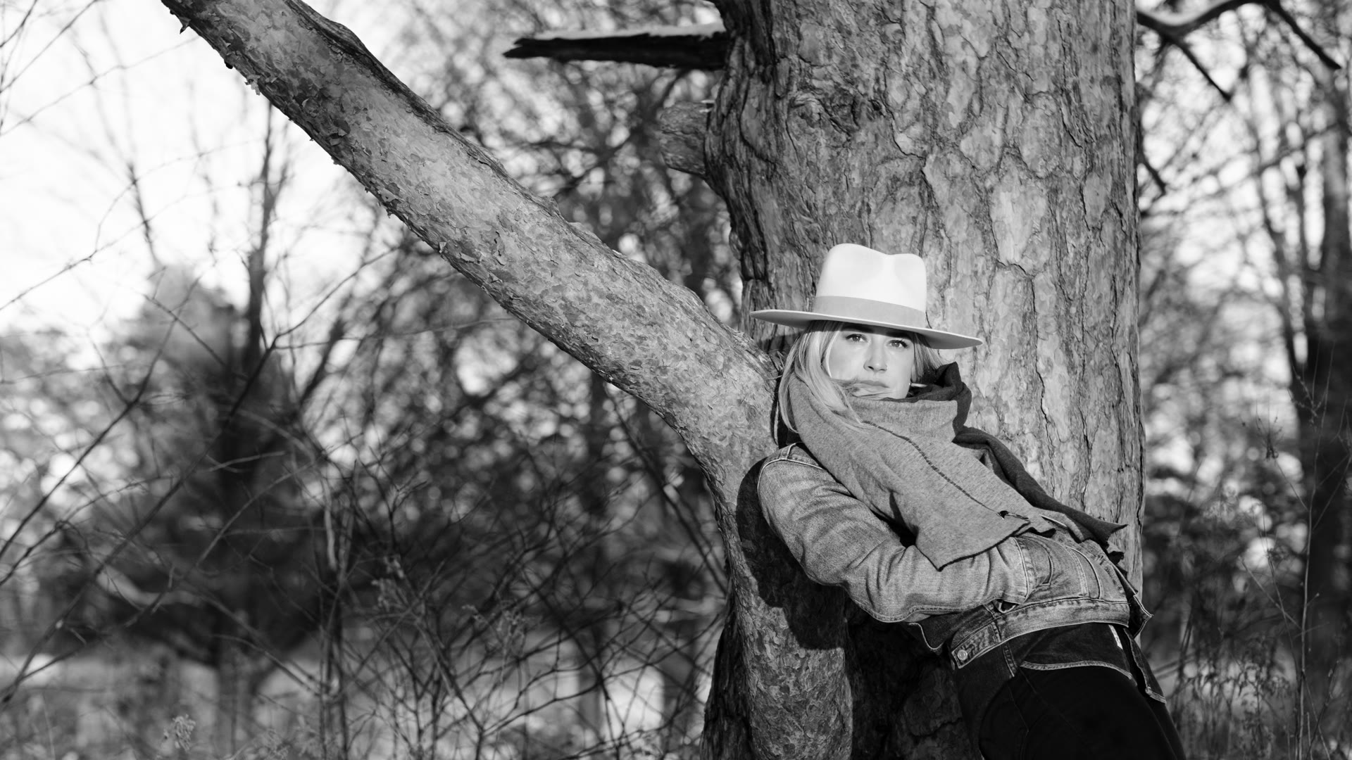 Portrait of Angelia Hughes in a wide-brimmed hat, leaning against a tree in winter woodland.